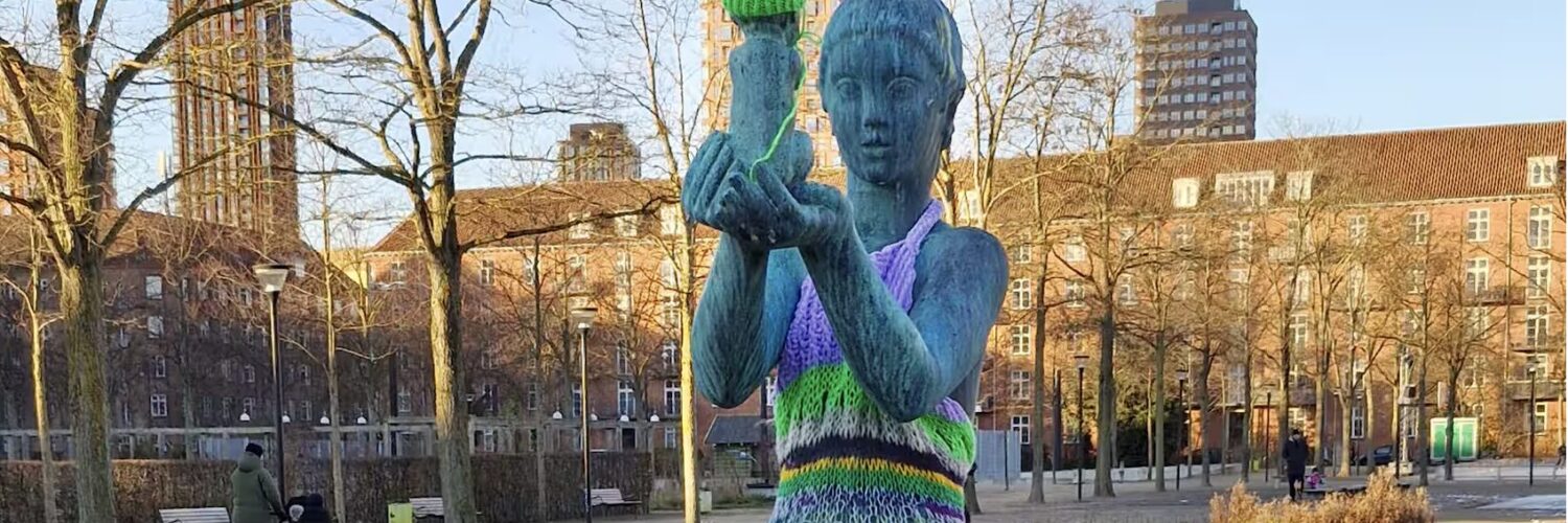 Statue de femme nue portant un enfant, habillée d’une robe rayée en tricot coloré et d’un bonnet vert, dans un parc à Copenhague.