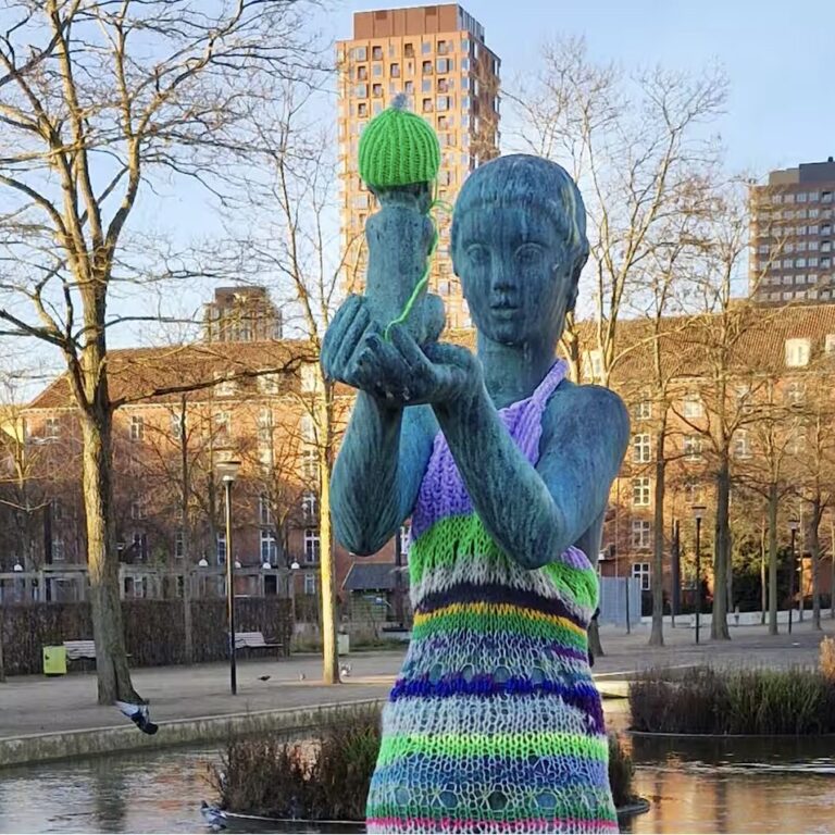 Statue de femme nue portant un enfant, habillée d’une robe rayée en tricot coloré et d’un bonnet vert, dans un parc à Copenhague.