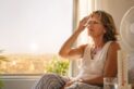 Femme de plus de 50 ans se rafraîchissant près d’une fenêtre pendant un épisode de fortes chaleurs, avec un ventilateur et un thermomètre en arrière-plan.